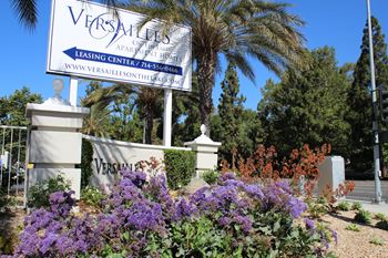 A blue and white sign for Versailles Apartments sits in front of a garden.